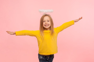 Wall Mural - Beautiful angelic little girl with halo over head raising hands as if flying and looking at camera with toothy smile, concept of kind obedient child. indoor studio shot isolated on pink background