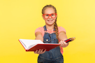 Portrait of clever creative little girl in red glasses holding notebook and pencils, school child smiling at camera showing homework, education concept. studio shot isolated on yellow background