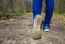 Young Girl In Jeans And Sneakers On Her Feet, Walks On The Road In The Green Forest In The Sunshine Outdoors In The Spring Time 