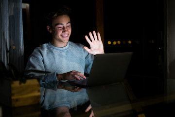 one caucasian man or teenager waving at someone doing videocall with someone in quarantine and lockdown - studying and doing homeowrk or working at night at home in videocall with his class online