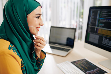 Wall Mural - cheerful smiling muslim young woman wearing earphones, programming on computer and talking to cowork