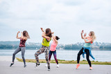 A group of young women, wearing colorful sports outfits, doing zumba exercises outside by city lake. Dancing training to loose weight in summer. Healthy lifestyle concept. Female sport leisure.