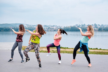 A group of young women, wearing colorful sports outfits, doing zumba exercises outside by city lake. Dancing training to loose weight in summer. Healthy lifestyle concept. Female sport leisure.