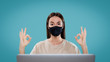 © South House Studio - A portrait shot of cute brunette female works isolated with her computer via freelance, she wear mask and shows ok signs with both hands
