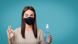 © South House Studio - Woman in medical mask holds a sanitizer and shows ok sign, isolated over blue