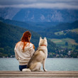 © voltgroup - A young woman with brown hair and white sweater is sitting at the pier at the lake with calm water. A Siberian husky female dog is lying down near the girl. The Alps mountains in the background.