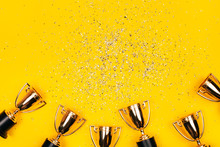 Golden Winner Cups With Silver Sparkles On A Yellow Background With Copy Space. Festive Concept. Flat Lay Style.