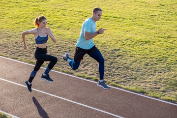 Wall Mural - Young couple of fit sportsmen boy and girl running while doing exercise on red tracks of public stadium outdoors.