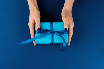 Wall Mural - Woman holding gift box with ribbon against classic blue background, top view