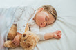 © kaloriya - Toddler baby sleeping on white sheets hugging soft brown teddy bear. Children's nap time or bedtime. Child sleep deprivation. Child sleep routine and schedule.