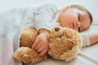 © kaloriya - Toddler baby sleeping on white sheets hugging soft brown teddy bear. Children's nap time or bedtime. Child sleep deprivation. Child sleep routine and schedule.  Close up.