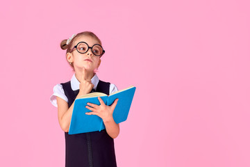 Wall Mural - primary school girl in uniform, round glasses without lenses holds a notebook in her hands with a thoughtful emotion on her face, posing on a pink background in the studio.