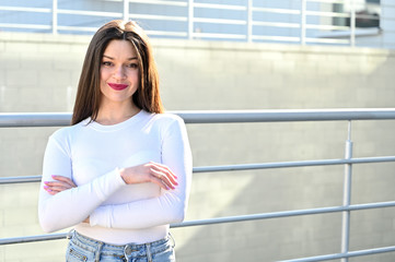 Wall Mural - Caucasian portrait of a pretty brunette girl in a white blouse and blue jeans stands near the railing with a smile against the background of a gray wall of a building on a sunny spring day.