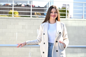 Wall Mural - Caucasian portrait of a lovely brunette girl in a light coat and blue jeans with a smile stands near the railing on a sunny spring day.