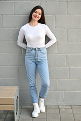 Wall Mural - Vertical Caucasian portrait of a pretty brunette girl in a white blouse and blue jeans stands near the wall of a building on a spring day in the city. Model is happy to pose.