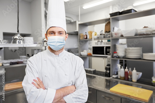 health, safety and pandemic concept - male chef cook in toque with crossed hands wearing face protective medical mask for protection from virus disease at restaurant kitchen