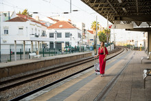 
Beautiful Woman At The Train Station. A Young Woman Dressed In A Red Suit With A Suitcase Walks To Her Train Station. In A Good Mood With A Smile On His Face.