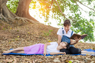 Canvas Print - Cute two children girl sitting under big linden tree reading book and enjoy in beautiful summer park.