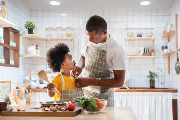 Wall Mural - Happy family with African father and son dress up together before cooking in the white kitchen. Single Dad Chef with black kid helper in yellow shirt preparing food and looking at Each Other at home. 