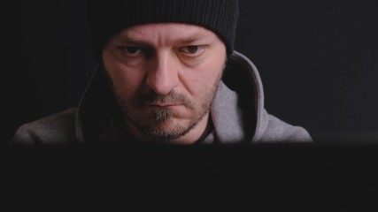 Serous man with black cap and hoodie sitting behind the computer monitor in the dark, typing, focused on screen, hacking computer system concept