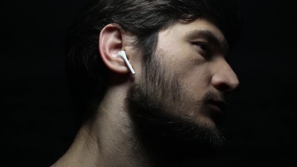 Wall Mural - Dark close-up portrait of young man with wireless earphones on black background.