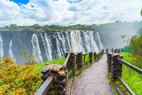 Fototapeta  - Walking way with view to the dramatic clouds and waterfall at Victoria Falls on the Zambezi River, Zimbabwe, Zambia.
