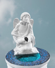 White statue of an angel by the bowl of holy water on his knees with a jug in his hands and an Orthodox cross isolated against a blue sky