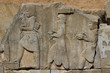 starożytne płaskorzeźby wykute w blokach kamiennych przedstawiające  mężczyzn idących z darami na jednej ze ścian w persepolis w iranie