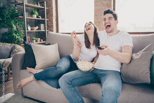 Portrait of nice attractive cheerful cheery couple sitting on divan watching comedian hilarious show comedy having fun quarantine at modern industrial loft style brick interior living-room flat