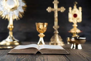 Wall Mural - Catholic religion concept. Catholic symbols composition. The Cross, monstrance,  Holy Bible and golden chalice on wooden altar and gray background. 