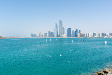 Abu Dhabi, United Arab Emirates skyline with a bright blue sky, sailboats, seagulls and landmark buildings in background. Luxury travel destination.