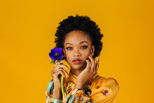 Portrait Of An Elegant  Young Woman Holding A Blue Flower With Orange Finger Nails, Isolated On Yellow Background