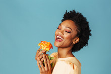 Portrait Of A Happy Young Woman Holding Yellow Orange Rose And Smiling With Eyes Closed, Isolated On Blue Background