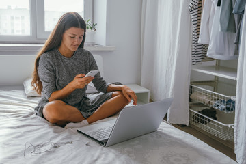 beautiful young woman with long dark hair wearing gray casual dress sitting at the bed and using smartphone and notebook, freelance concept