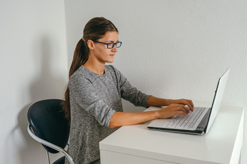 young beautiful woman wearing glasses and gray dress working with laptop against white wall