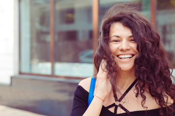 Happy face with curly hair looks at camera, smiles, adjusting her hair with right hand. Outdoors