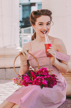 Portrait Of Happy Beautiful Girl In Pink Retro Dress And Pin Up Hairstyle Drink Lemonade And Smile Outdoors. Attractive Fashionable Young Vintage Woman Posing With Flowers Basket And Lemonade In Park