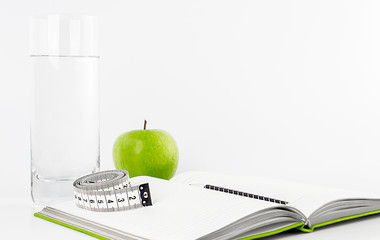 Open empty daily, pen, measuring tape, cup of water, fresh green apple on a light background. The concept of a healthy lifestyle, health diary. Copy space. 