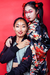 two pretty geisha girls friends: modern asian woman and traditional wearing kimono posing cheerful on red background