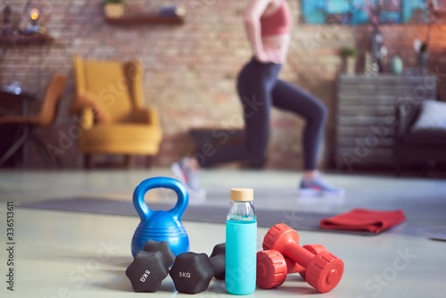 Exercising at home in living room. Focus on fitness equipments, barbell and kettlebell. Woman doing walking lunges exercise in the background. Concepts about home workout, fitness, sport and health.