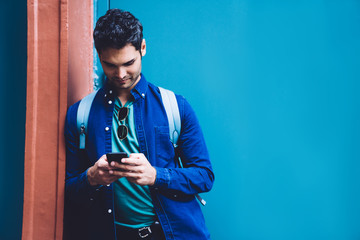 handsome modern man in trendy shirt leaning on blue wall on street and using mobile phone smilingly,