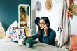 Young charming stylish woman in a home office sits at a desk, looks at a laptop and speaks while smiling on the phone. The concept of freelance and remote work. Stay safe and self-isolation.