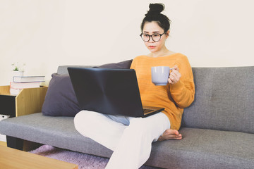 Young beautiful asian woman wore orange knitwear, sitting on gray sofa ,work online lifestyle,relax at home with coffee mug,home working laptop,work from home