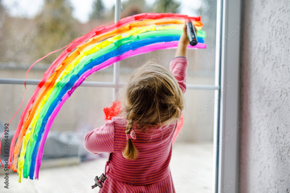 Adoralbe little toddler girl with rainbow painted with colorful window color during pandemic coronavirus quarantine. Child painting rainbows around the world with the words Let's all be well. - obrazy, fototapety, plakaty 