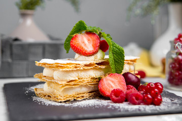 Wall Mural - Closeup on a strawberry and whipped cream mille-feuille with fresh berries on a black stone board on the white background, horizontal