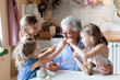 Kids treat grandmother at home. Happy family eating cookies in cozy kitchen. Senior woman and funny children tasting delicious food together, enjoying handmade pastries.