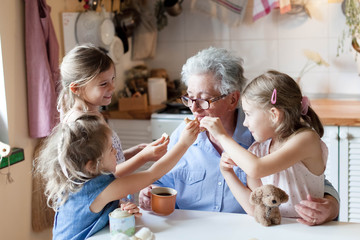 Wall Mural - Kids treat grandmother at home. Happy family eating cookies in cozy kitchen. Senior woman and funny children tasting delicious food together, enjoying handmade pastries.