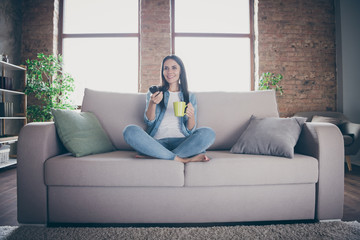 Wall Mural - Full body photo of cheerful cute girl have covid-19 quarantine rest relax free time sit divan legs crossed watch tv switch channels enjoy hold cacao coffee cup in house indoors