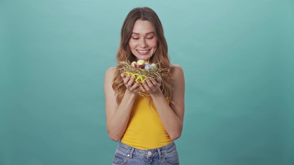 Poster - Happy young girl shows a nest with Easter eggs to the camera isolated on blue background