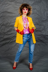 Wall Mural - Girl in a yellow jacket and blue jeans with an afro hairstyle. Fashion of the eighties, disco era. Studio photo on gray background.